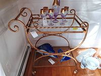 Brass rolling bar cart with two glass shelves and a curved scrollwork design. Top shelf has holders for bottles. Contents on the cart including glassware and blue glass decor are not included.