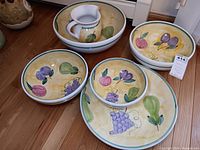 Overall view of five ceramic nesting bowls, a ceramic pitcher, and the large platter all painted with fruit designs and yellow background.