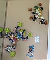 Three ceramic tree frogs of various sizes mounted on a beige wall. Largest has green palms and colorful body; medium frog is green-black with orange and yellow details; smallest is blue with dots and flowers.