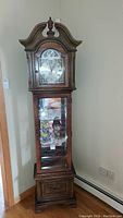 Front view of the wooden grandfather clock and curio cabinet showing clock face, glass display with items inside, and base drawer.