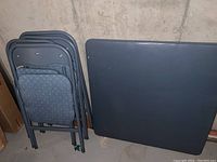 Stacked folding chairs with blue patterned upholstery next to folded blue folding table top with visible tear.
