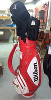 Red and white Wilson golf bag with multiple golf clubs inserted and black protective headcovers in a basement environment.