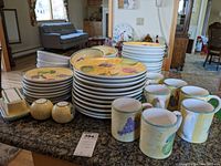 Photo of stacked dinner plates, luncheon plates, bowls, mugs, butter dish, salt & pepper shakers with colorful fruit patterns on yellow background.