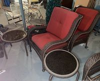 Two brown wicker outdoor chairs with red cushions and two round side tables placed on basement floor with other furniture in background
