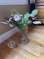 Porcelain roses in glass vase with red, pink, and yellow roses and green leaves positioned on hardwood floor next to wall.