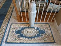 Photo showing the Persian throw rug with floral design and the tall white lidded vase on top.