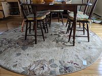 Round beige floral patterned rug under a wooden dining table with chairs, showing the rug's full design and size.