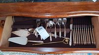 Wide view of flatware drawer showing knives, forks, spoons and serving utensils all silverplated with wood or dark colored handles along the right side and center, brass candle snuffer on left side