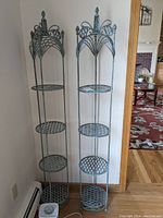Two tall wrought iron 4-tier plant stands with lattice shelves, seen from front in a corner of a room with wooden floor and wall baseboard heater.