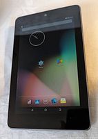 Front view of Asus Nexus 7 tablet displaying home screen with app icons and clock on a colored gradient background.