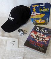 Full view of lot on white fabric background showing all four items: black cap, Hot Wheels Yellow Submarine toy in package, drum-shaped keychain, and DVD.