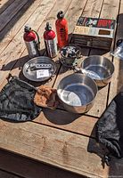 Shows full kit of items including stove, fuel bottles, pots, heat shield, gloves, and mesh bags laid on wooden surface.