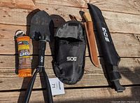 Full lot displayed on wooden surface including collapsible shovel, machete with sheath, fillet knife with sheath, and paracord.