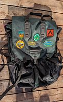Front view of the Eureka Portage 115 backpack showing green flap with patches and black mesh lower half with straps.