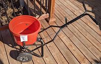 Watercraft garden spreader showing orange hopper, black frame, wheels, and handle from rear angle on wooden deck.