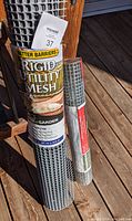 Two rolls of rolled metal mesh leaning against a wooden post on a deck floor. One roll is labeled 'Better Barriers Rigid Utility Mesh', the other is a smaller roll with a red label identified as welded hardware mesh.