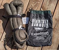 Hennessy Hammock folded with suspension straps secured and storage bag labeled 'Hennessy Hammock Expedition Asym Zip' on wooden floor.