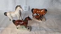 Three horse figurines displayed on white cloth backdrop: cream horse, brown horse, dark brown horse with foal.