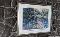 Framed watercolor print showing pintail ducks in a pond with surrounding foliage and reflections. The frame is silver-tone with a white mat and a black double inset line. Wall stone background visible.