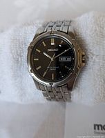 Front close-up showing watch face details including black dial, day and date window, and stainless steel case and bracelet.