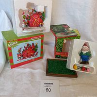 Photo showing The Grinch figurine kit partially removed from styrofoam with its original box and the Gnome & Garden kit pieces, including gnome figurine, artificial grass base, and flower bouquet on a white cloth.