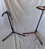 Pair of guitar stands side by side on a white cloth background. One stand is missing the bottom guitar support fork.