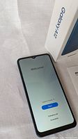 Samsung Galaxy A12 smartphone powered on showing the welcome screen. Device in black color. Original box visible in background.