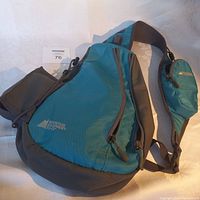 Front and side view of blue and black Mountain Equipment Co-op sling pack showing zippered main compartment and integral small pouch on strap.
