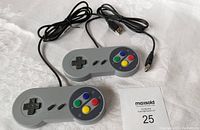 Photo showing two grey USB wired gaming controllers styled like classic Super Nintendo controllers placed on white fabric background with MaxSold tag.
