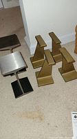 Six I-beam shaped metal bookends arranged on carpet. Four brass toned with angled tops, two silver steel finish with flat tops.