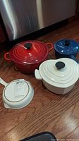 All four pieces together on wood floor: white skillet, white pot with lid, red pot with lid, blue saucepan with lid
