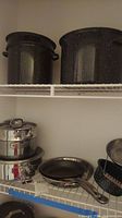 Shelf with large black speckled enamel pots and stainless steel cookware including skillet