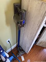 Dyson DC44 Animal cordless vacuum upright on floor beside wall with charging dock and cord attached, blue cleaning wand and head visible