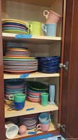 Shelving unit with multiple stacks of colorful Fiesta plates, bowls, cups, and shakers arranged by type and color.
