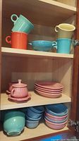 Three shelves in wooden cabinet holding various Fiesta brand cups, bowls, plates, and cream and sugar set in multiple colors
