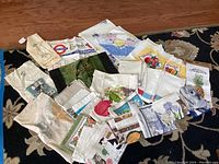Overview of assorted tea towels, table cloths and bags arranged on carpet showing colorful prints including cats, fruits, and flowers.