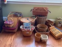 Twenty-one assorted Longaberger baskets in various sizes and styles are displayed on a hardwood floor against a green wall. Several of the baskets have cloth liners in different colors and patterns. A large round basket with a lid is visible, along with several rectangular and oval baskets. The box of basket ornaments and a screensaver disc are mentioned but not visible in detail.
