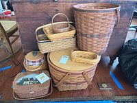 Overview of eight baskets of varied sizes and shapes, some with lids and plastic liners, arranged on a floor in front of wooden furniture.