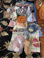 Wide photo showing assorted colorful and patterned linens including placemats, basket liners, and tie ons spread out on a carpet with a wooden basket to the right side.