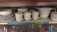 View of plates, bowls, mugs, and pitcher arranged on cabinet shelf.
