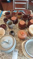 Full lot of approximately 28 pottery and redware items on granite countertop including cups, bowls, candle holder, planter with lid, arranged clustered together.