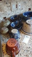 Photo showing 8 handmade pottery pieces including bowls and strainers with varying hole configurations and glaze colors, placed on a kitchen counter.