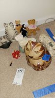 Wide image showing pottery cat figurine, several teddy bears, a white dog figure, and other small animal figures arranged on carpet.