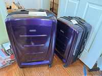 Photo showing two purple Samsonite suitcases of different sizes standing on wooden floor near a white door.