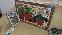 Two stained glass panels displayed on a couch: a large 21x33 inches floral panel with potted plants and a smaller 13x10 inches floral panel with purple petals.