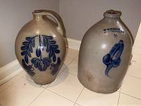 Two salt glazed stoneware jugs side by side on tiled floor showing overall form and decorations.