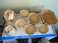 Eight stoneware molds arranged on a wooden shelf showing various shapes and sizes, including bundt and floral style molds.