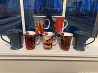 Front view of all seven mugs showing two solid blue and five decorated mugs in various sizes and colors