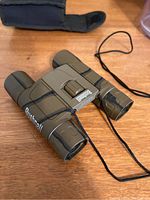 Front top view of Bushnell camo binoculars with neck strap, case in background.