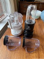 Small white Cuisinart food processor alongside black Magic Bullet single shot blender with clear blending cup attached, and two Magic Bullet travel cups with lids placed on wooden table.